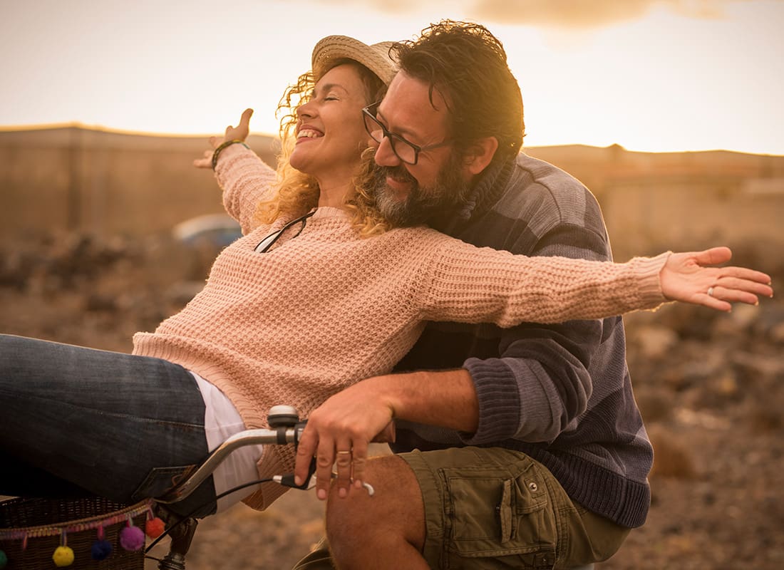 About Our Agency - Cheerful Middle Aged Couple Having Fun Spending Time Together While Riding a Bike Together in the Countryside at Sunset