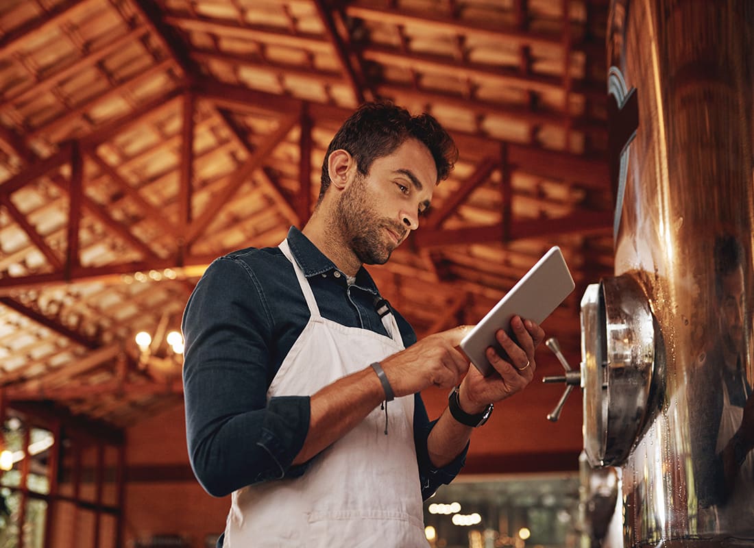Business Insurance - Portrait of a Young Male Brewery Owner Using a Tablet While Standing Next to Beer Brewing Equipment