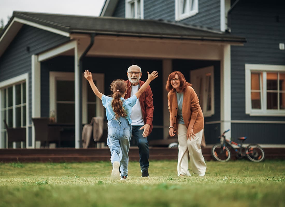 Personal Insurance - View of a Young Girl Running Across the Grass Toward her Smiling Grandparents Standing in Front of Their Two Story Home