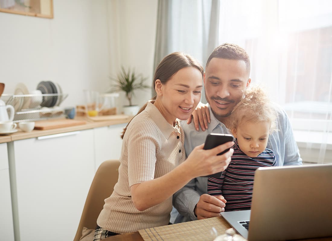 Read Our Reviews - Cheerful Young Diverse Parents Sitting in Front of the Computer in the Kitchen with Their Young Daughter While Looking at a Phone