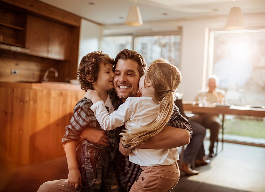 We Are Independent - Cheerful Young Father Hugging his Son and Daughter in the Kitchen with Warm Light Shining Through the Windows in the Background